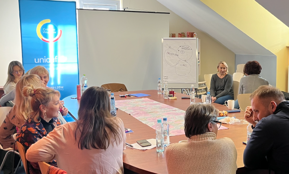 Grupa osób podczas warsztatów przy stole konferencyjnym, w tle baner UNICEF i tablica z notatkami